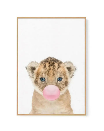 Framed canvas art print featuring a young lion cub with tan and brown striped fur and bright blue eyes, blowing a large bubble gum bubble in soft pink. The cub's head is centered against a clean white background. The illustration has a soft, watercolor-like quality. The canvas is mounted in a natural wood frame with a warm honey tone.