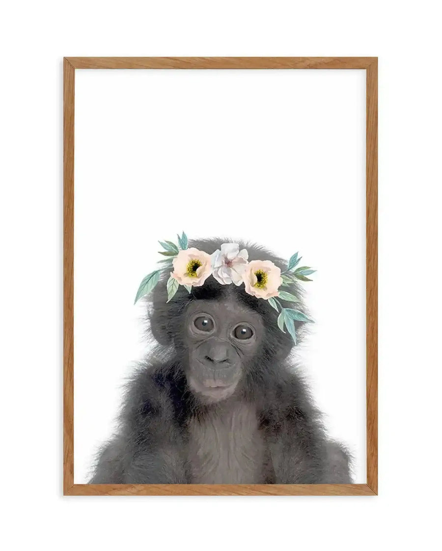 Watercolour art print featuring a baby chimpanzee with dark grey fur and large dark eyes, wearing a flower crown with pale pink, cream, and white blossoms and light green leaves, displayed in a natural oak frame with white border.