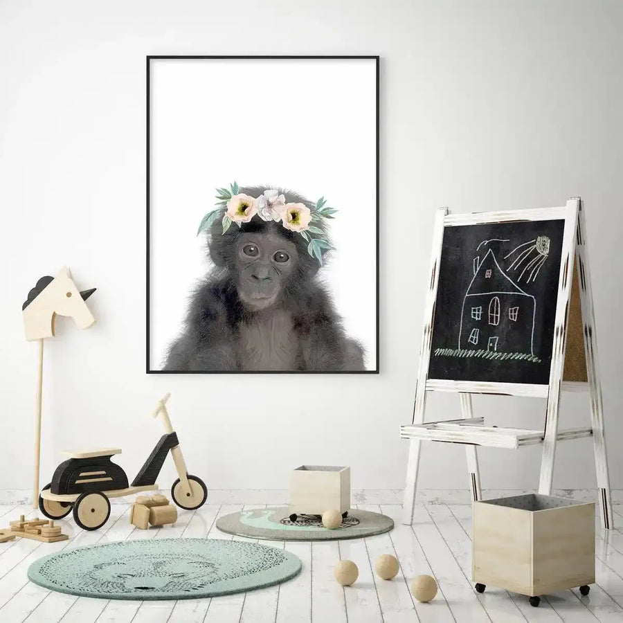 Lifestyle shot of a child's playroom featuring a framed art print of a baby chimpanzee wearing a flower crown. The print is displayed on a white wall above a wooden toy unicorn and a wooden balance bike. The chimpanzee has dark fur and large, expressive eyes, with a crown of pale yellow and pink flowers with green leaves. The room has white wooden floorboards, two round rugs in muted green and grey, a small wooden box on wheels, and a white easel with a chalk drawing of a house.