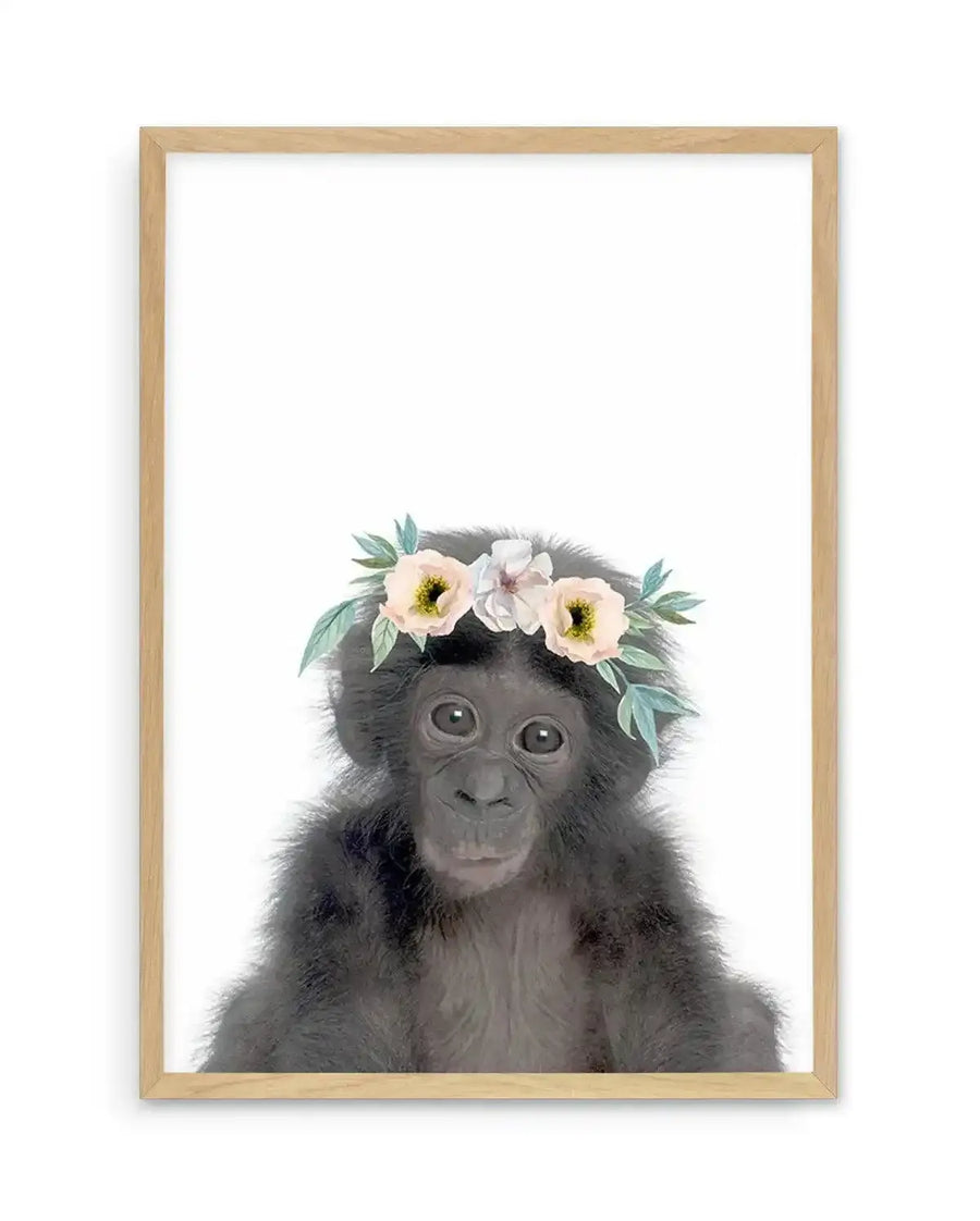 Watercolour art print featuring a baby chimpanzee with dark grey fur and large, expressive eyes, wearing a delicate flower crown with pale pink and white blooms and soft green leaves, displayed in a natural oak frame with white border.