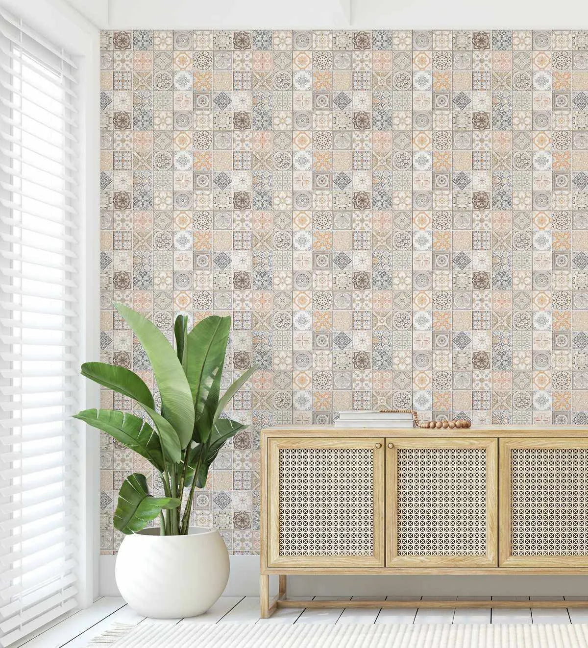 Lifestyle shot of a room featuring Little Moroccan Tiles Wallpaper. The wallpaper covers a full wall and displays a repeating pattern of small square tiles, each with a unique geometric or floral design in muted tones of beige, cream, light grey, soft peach, and subtle brown. The wallpaper has a textured, aged appearance. In the foreground, a large green plant in a white pot sits next to a window with white blinds. A light wood cabinet with woven cane doors is positioned against the wallpapered wall.