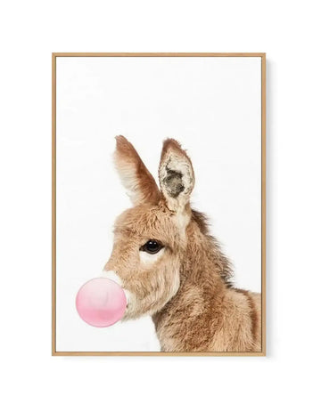 Framed canvas art print featuring a tan and white llama with long ears against a white background, blowing a bright pink bubble. The llama's face is rendered in detailed, photorealistic style with a large dark eye visible. The canvas is mounted in a natural wood frame with a white border.