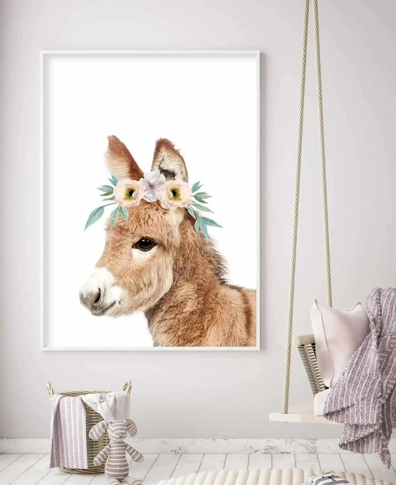 Lifestyle shot of a nursery featuring a framed art print of a baby donkey wearing a flower crown, displayed on a white wall above a woven basket with blankets and a striped bunny toy. The art print features a close-up profile of a light brown donkey foal with dark eyes and a white muzzle, adorned with a watercolor flower crown of pale pink and white blossoms with green leaves. A light wood swing with a pink pillow and striped blanket hangs to the right.