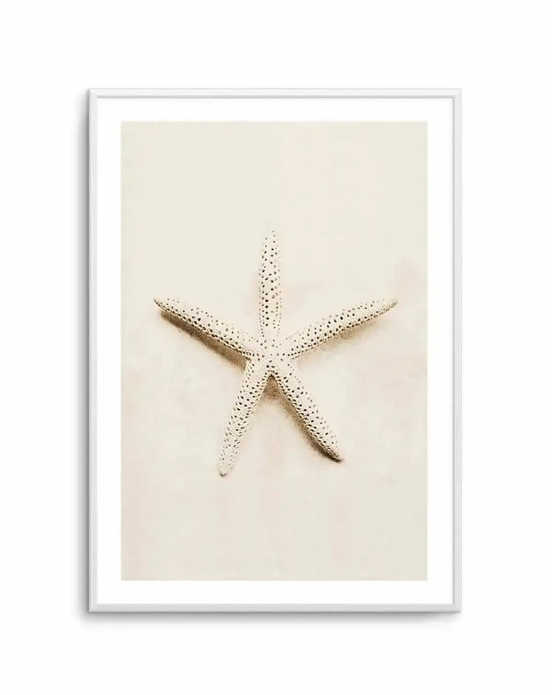 Framed photography art print featuring a white starfish with five arms, speckled with small black dots, lying on a light beige sandy background, with a white frame and white border.
