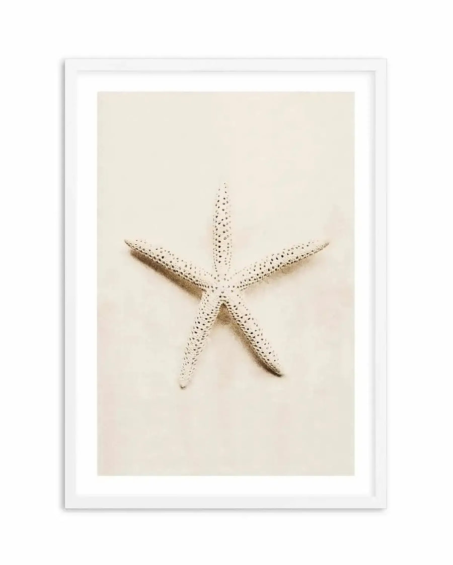Framed photography art print of a single starfish on sand, displayed in a white frame with white border. The starfish is light cream with dark speckles, positioned diagonally on a soft, light brown sandy background.