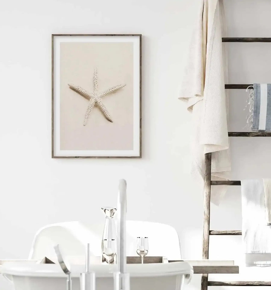 Bathroom:
Framed art print of a white starfish displayed on a light cream background, with natural wood frame and white border, hanging on a white wall in a bright, minimalist bathroom. A rustic wooden ladder with white towels draped over it is visible on the right, and a white bathtub with a chrome faucet and decorative items is in the foreground.