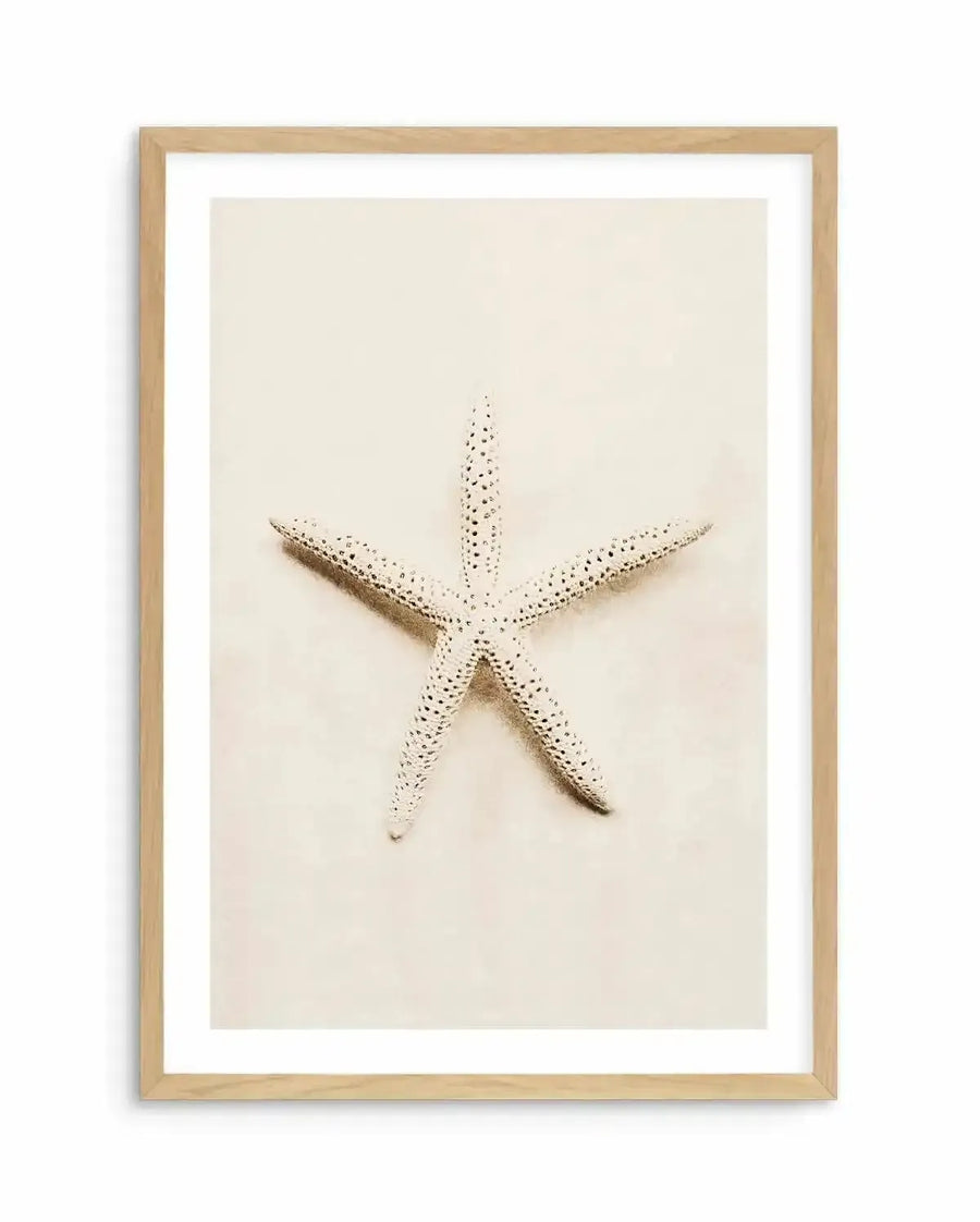 Framed photography art print featuring a single starfish on a sandy background, with natural wood frame and white border. The starfish is light in colour with small dark spots and is positioned diagonally across the frame, casting a soft shadow on the light beige sand.