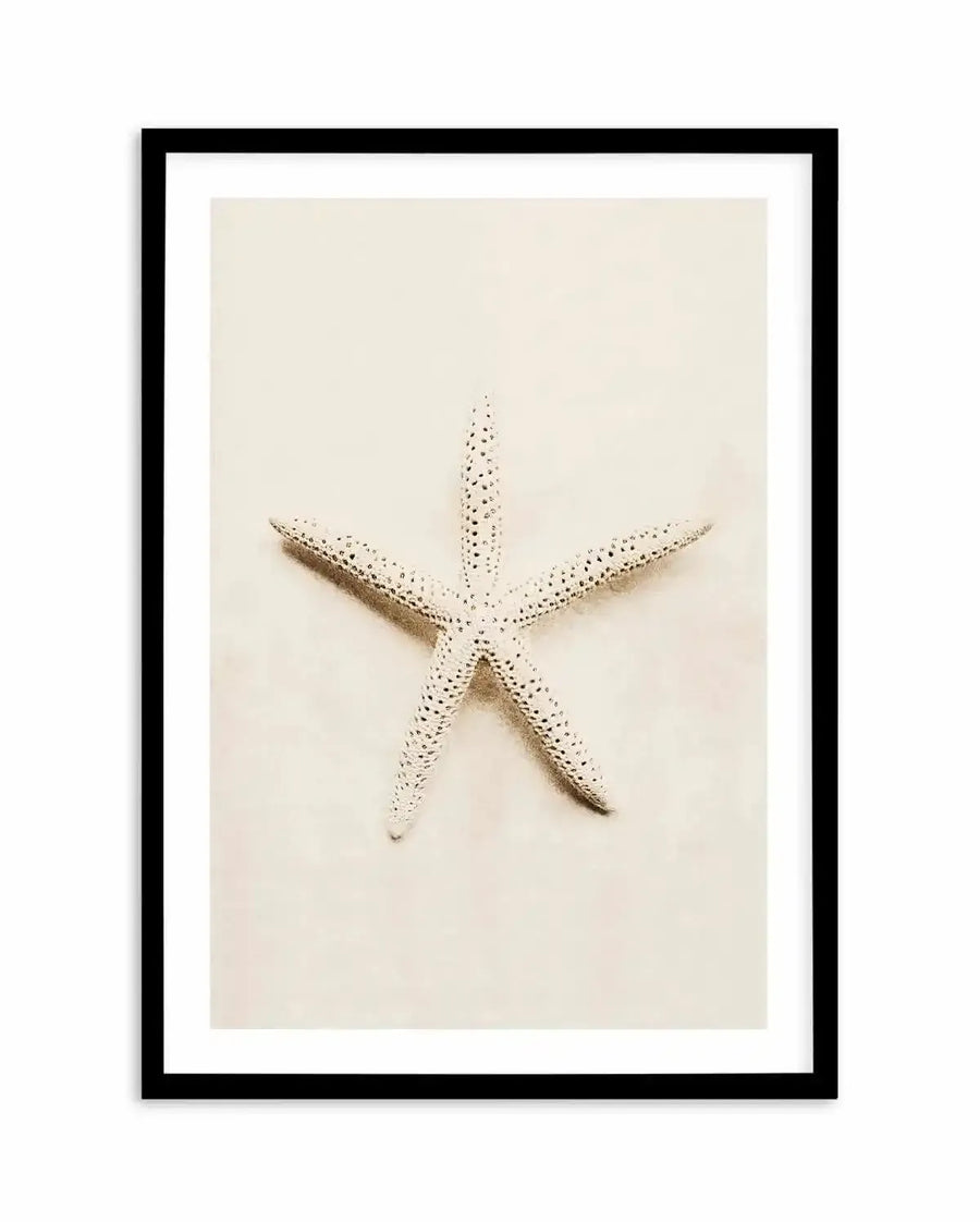 Framed photography art print featuring a single starfish centered on a light, sandy background, displayed in a black frame with a white border. The starfish is light-coloured with small, dark speckles covering its surface, casting a subtle shadow on the textured sand.