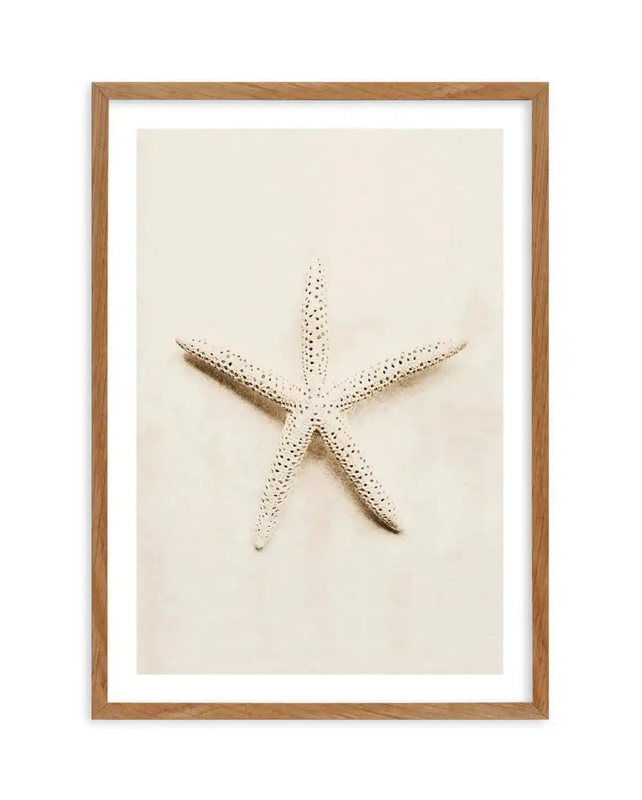 Framed photography art print featuring a white starfish with textured bumps, centered on a light beige background, displayed in a natural oak frame with white border.