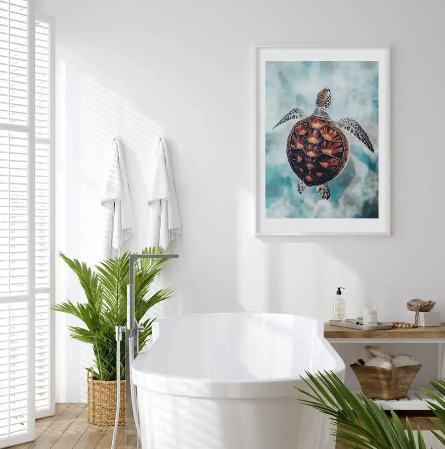 Lifestyle shot of a minimalist bathroom with a framed art print of a sea turtle swimming in turquoise water. The print features a brown and orange patterned sea turtle with speckled flippers, viewed from above, against a mottled blue and white background, displayed in a white frame with a white border. The bathroom includes a white freestanding bathtub, a large green plant in a woven basket, and a wooden shelf with bath products.