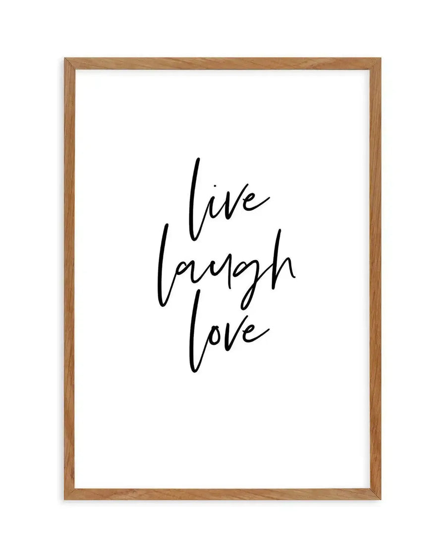 Framed art print with handwritten-style black cursive text reading "live laugh love" stacked vertically on a white background. Natural wood frame with warm tan finish. Simple, minimalist design with flowing script lettering centered in the frame.
