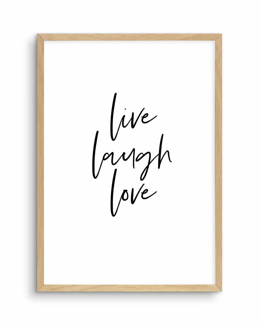 Framed art print with natural wood frame displaying handwritten-style black text on white background reading "live laugh love" in three stacked lines with flowing, cursive letterforms.