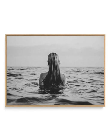 Framed black and white photograph of a woman with long wet hair, seen from behind, standing in the ocean with gentle waves around her, displayed in a natural oak frame with white border.