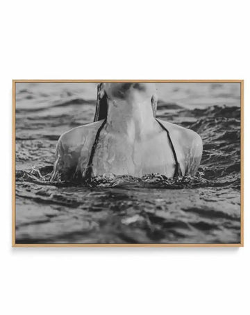 Black and white framed canvas art print titled L'Ocean II featuring a woman partially submerged in ocean waves, shot from shoulders up. The photograph captures water splashing around her neck and shoulders against a dramatic seascape background with rolling waves and distant shoreline. The image has a vintage film quality with grainy texture and high contrast. The canvas is mounted in a natural wood frame with a warm tan finish and displayed against a white background.