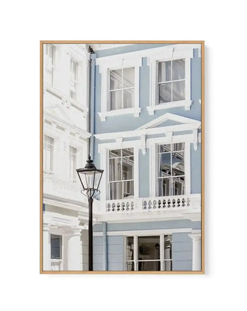 Framed canvas art print featuring a photograph of a light blue and white Parisian-style residential building facade. The composition shows three stories with multiple large windows with white frames and shutters, decorative white balustrade railings, and a black street lamp in the foreground. The image has a soft, minimalist aesthetic with cool blue-grey and white tones. The canvas is set in a natural wood frame with a white border, displayed against a white background.
