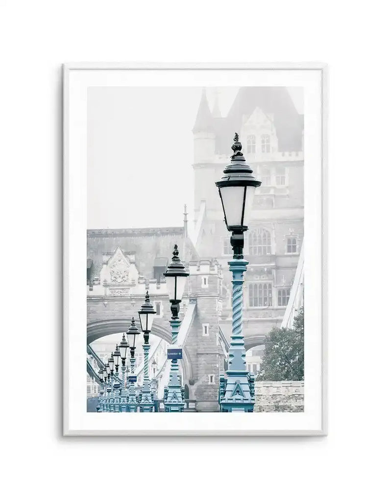 Framed photography art print of London's Tower Bridge on a misty day, with a row of ornate black and blue lampposts lining the bridge. The print has a cool, desaturated colour palette with soft blues, greys, and whites, and is displayed in a white frame with a white border.
