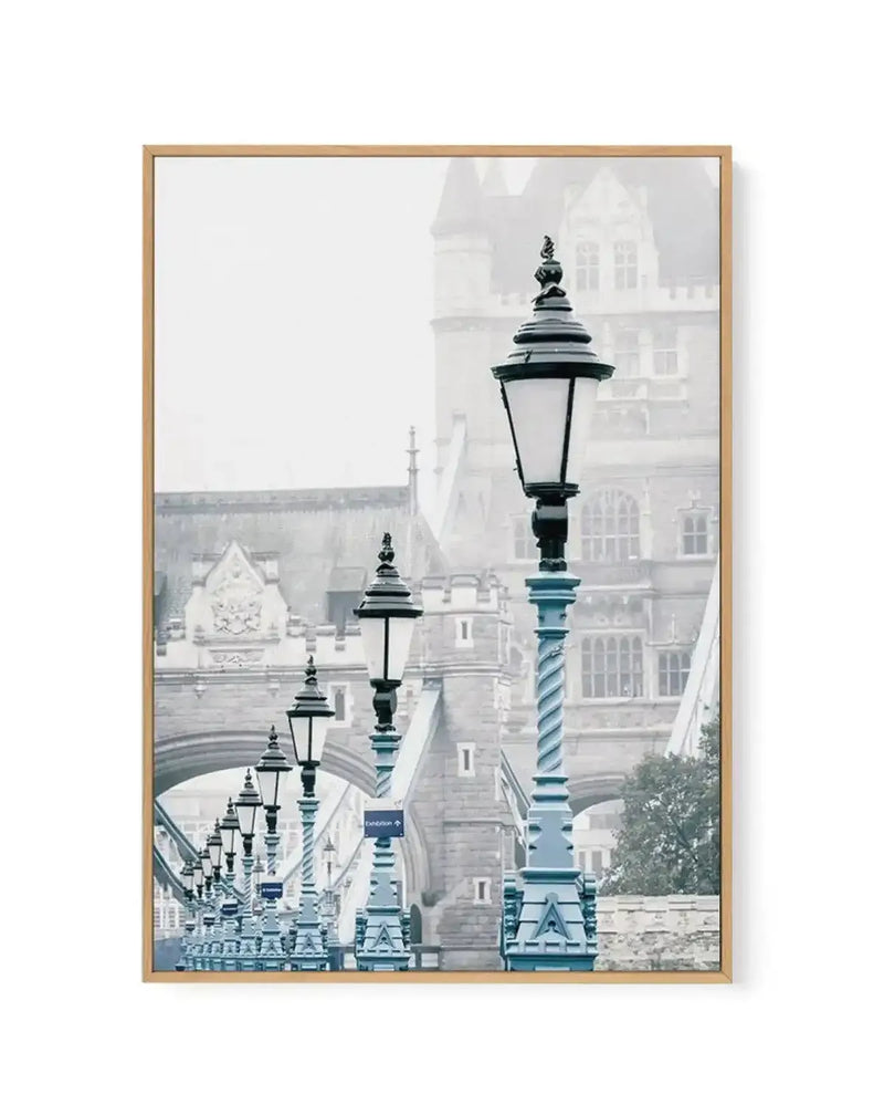 Framed canvas art print featuring a photograph of London street lamps in teal and dark green with ornate bases, arranged in receding perspective along a misty street. Tower Bridge and historic architecture blur in the foggy background. Natural wood-toned frame with white border surrounding the vertical composition.