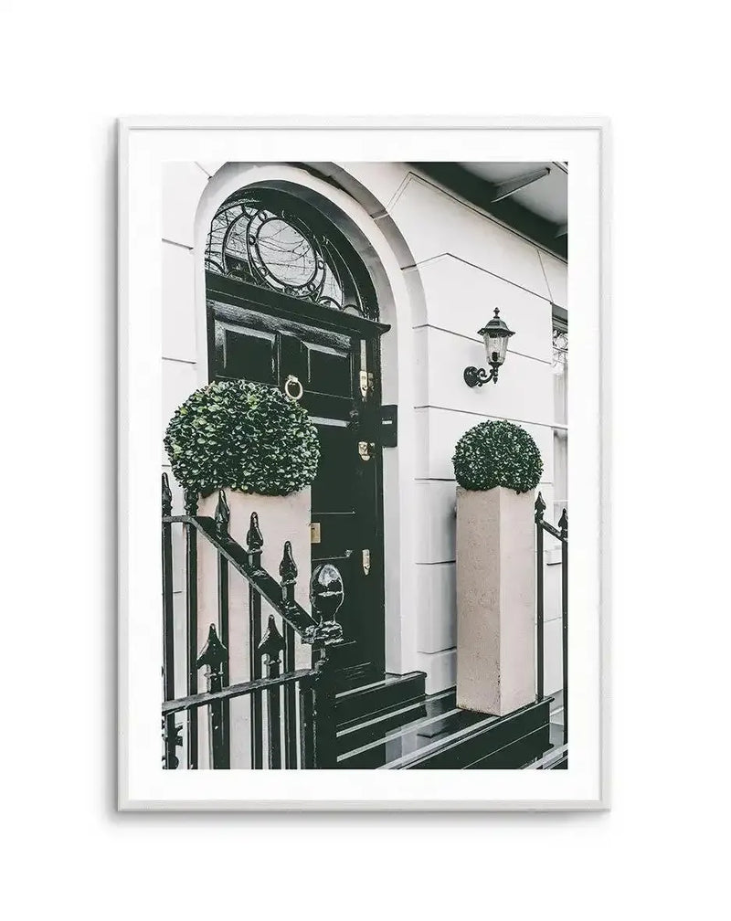 Framed black and white photograph of a classic London townhouse entrance with a black door, arched transom window, and two topiary plants in tall planters, with a black wrought iron fence in the foreground.