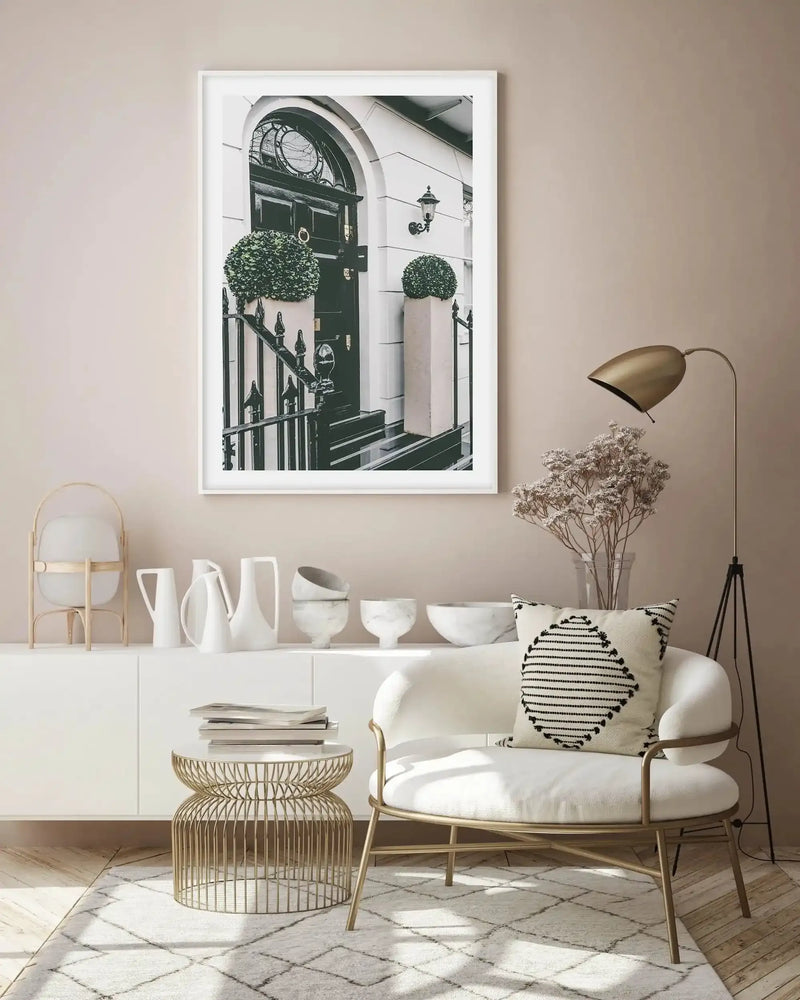 Large framed art print displayed above a white console table in a modern living room. The print is a black and white photograph of a classic London townhouse entrance, featuring a dark green door with a fanlight window above, flanked by black railings and two potted topiary plants. The room includes a white armchair with a black and white patterned cushion, a gold wire side table, and a gold floor lamp.