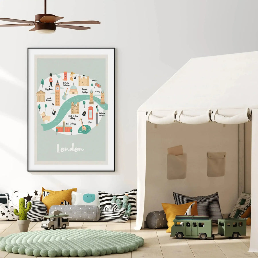 Lifestyle shot of a child's room featuring a framed art print of a London map illustration above a collection of floor pillows. The print, in a black frame with a white border, depicts a light blue map with a teal river, showcasing iconic London landmarks like Big Ben, the British Museum, Westminster Abbey, and Tower Bridge, along with whimsical elements like a red telephone booth and a guard. The room has light wood flooring, a light green textured rug, a white play tent, and various patterned pillows.