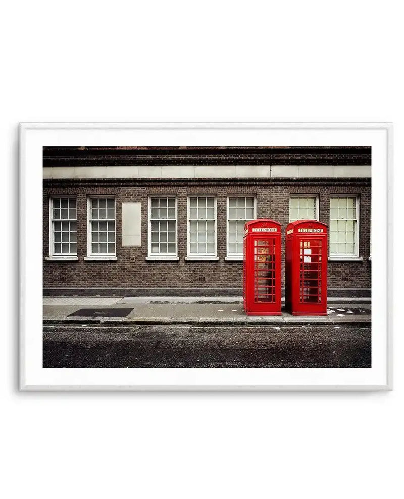 Framed photograph of two iconic red London phone booths standing side-by-side on a sidewalk in front of a brick building with several white-framed windows, with a white border and a white frame. The street in the foreground is dark and wet, suggesting recent rain.