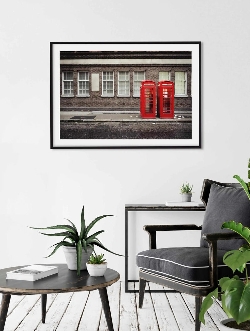 Lifestyle shot of a framed art print displayed on a white wall above a dark wooden side table and chair. The art print features a photograph of two iconic red London telephone booths standing on a sidewalk next to a brick building with white-framed windows, with a black frame and white border. The room includes a dark wooden round coffee table with a book and potted plants, and a dark grey upholstered chair with white piping.