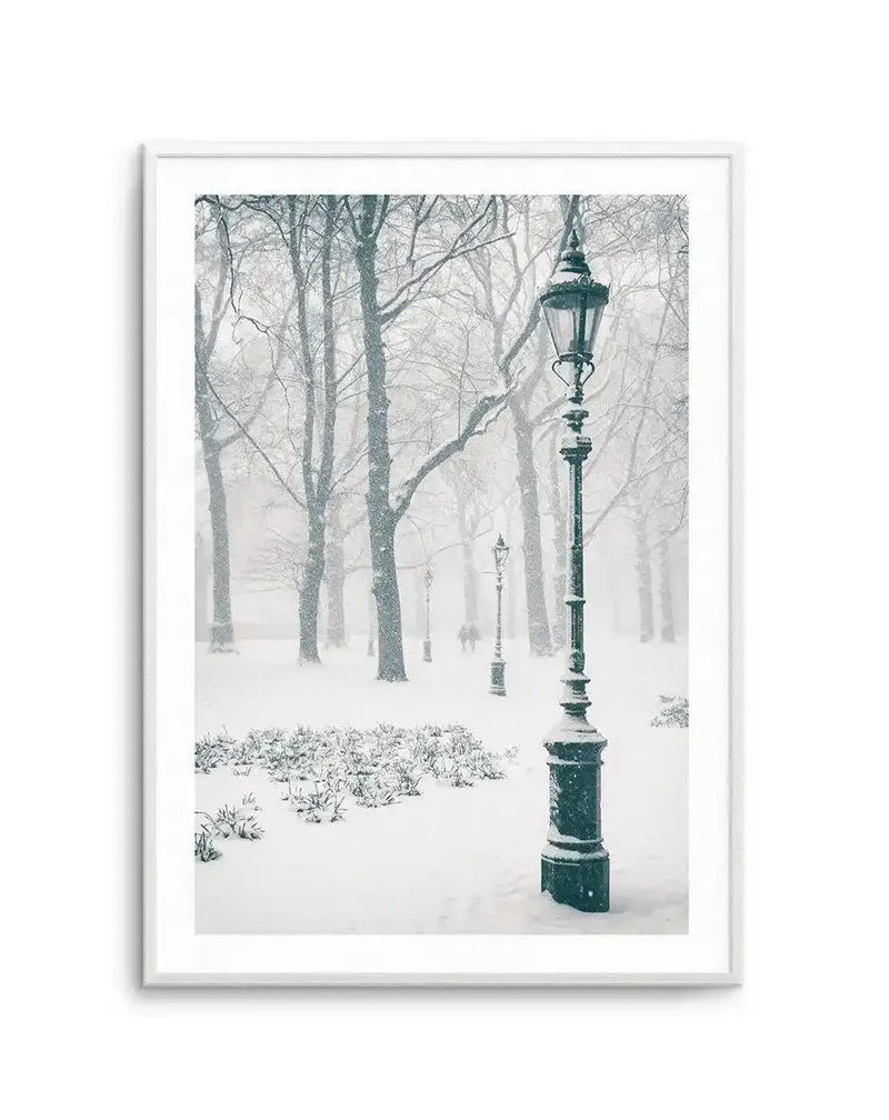 Framed black and white photograph of a snowy park scene with a white frame and white border. The composition features a tall, ornate black lamppost in the foreground on the right, covered in snow. In the background, bare trees are visible through falling snow, with several other lampposts and two figures walking in the distance. The ground is covered in a thick layer of snow, with some low bushes peeking through in the midground.