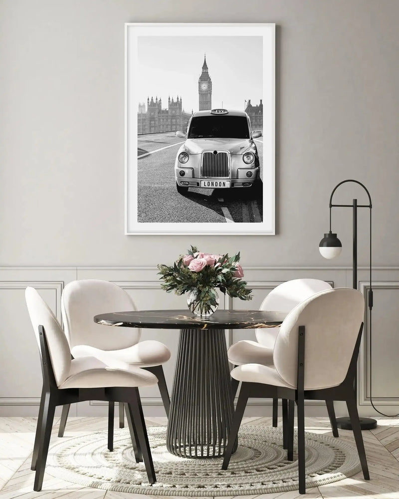 Lifestyle shot of a modern dining area with a large framed black and white photograph of a London taxi and Big Ben. The print is displayed above a black marble dining table with a ribbed black base, surrounded by four cream upholstered chairs with black legs. A vase of pink roses and greenery sits on the table. A black and white floor lamp stands to the right.