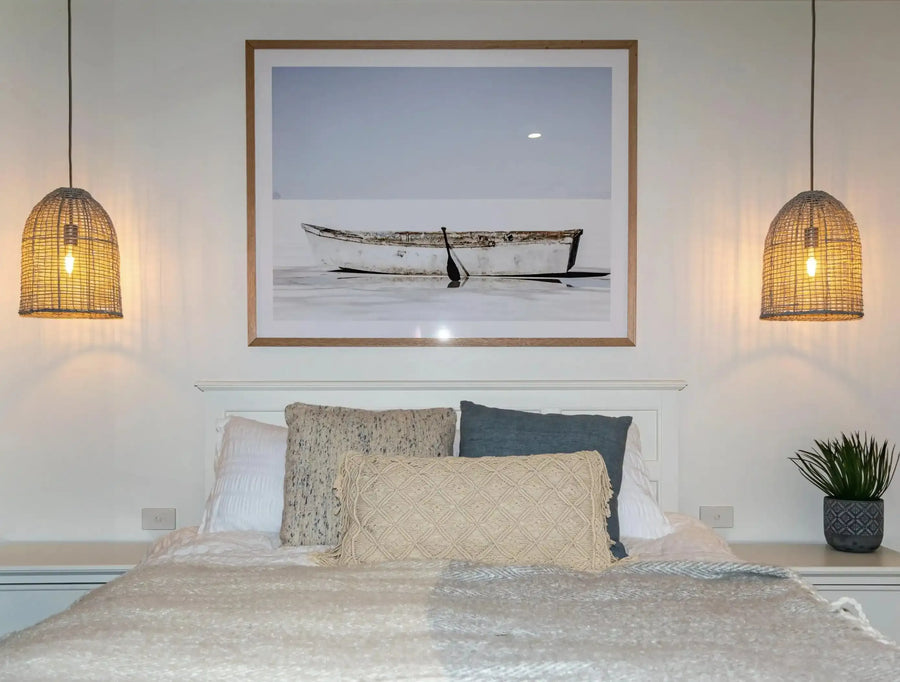 Large framed art print displayed above a white headboard in a neutral bedroom. The print features a weathered white rowboat with a single black oar resting in calm, shallow water under a pale blue sky, with a natural wood frame and white border. The bed is dressed with a cream textured throw and various pillows, and two woven pendant lights hang on either side of the bed.