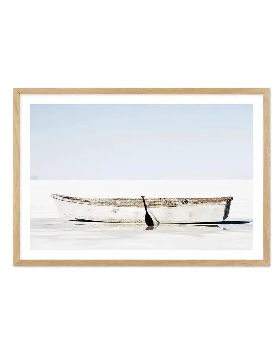 Framed photograph of a weathered white rowboat with a single black oar resting inside, floating on calm, reflective white water under a pale blue sky, with a natural wood frame and white border.