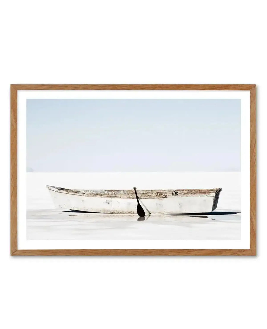 Framed photograph of a weathered white rowboat with a single oar resting in calm, shallow water under a soft blue sky, with a natural oak frame and white border.