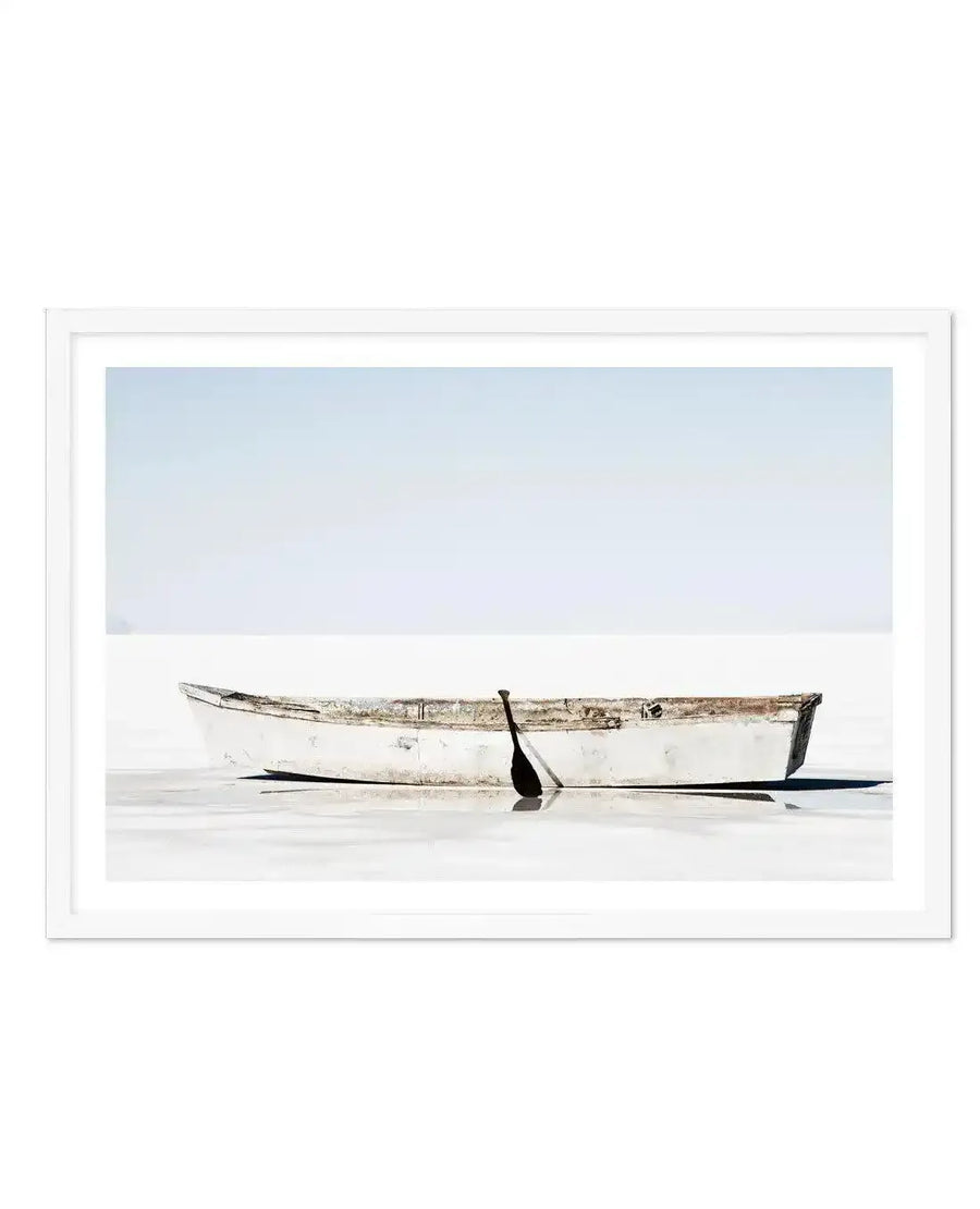 Framed photography art print of a minimalist coastal scene with a white wooden rowboat and a single black oar resting in shallow, still water, with a pale blue sky stretching above. The print is displayed in a white frame with a white border.