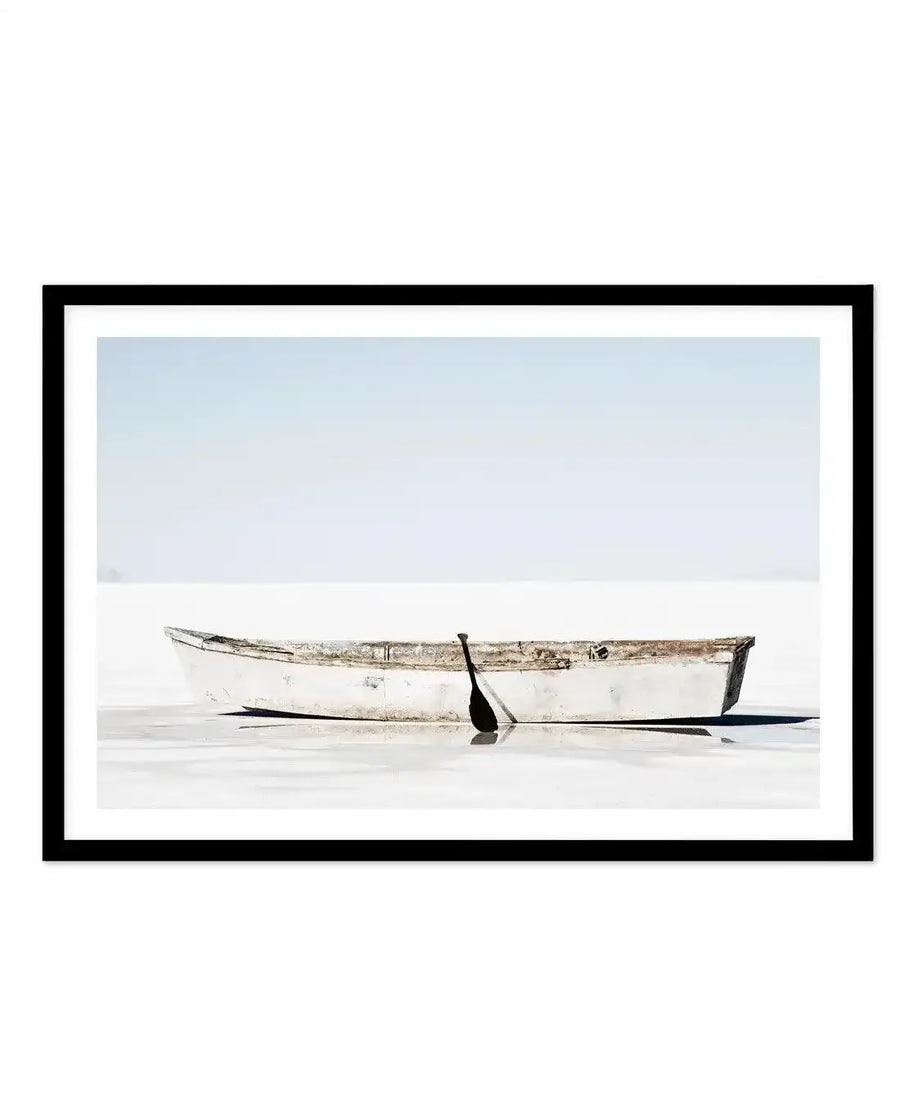 Framed black and white photograph of a weathered white rowboat with a single black oar resting in shallow, calm water, with a light blue sky stretching to the horizon. The print is displayed in a black frame with a white border.