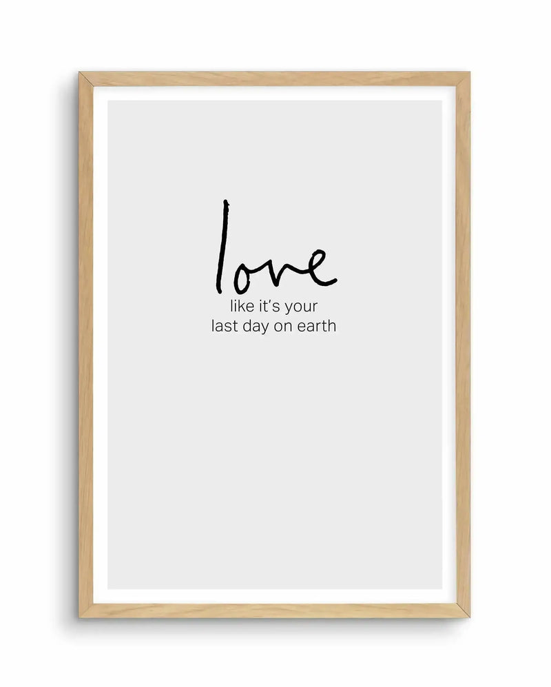 Art print with light wood frame and white border featuring handwritten-style black text on a light gray background. The word "love" appears in larger, casual script at the top, followed by smaller text reading "like it's your last day on earth" positioned below. The minimalist design uses simple black typography centered on the page, conveying an inspirational message with understated elegance.