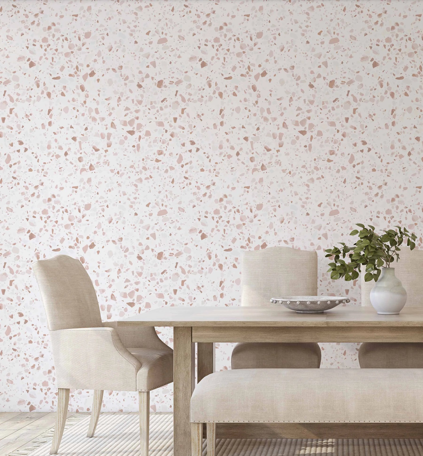 Lumiere Terrazzo Wallpaper displayed on a bedroom wall behind a light wood dining table set with cream upholstered chairs and bench. The wallpaper features a terrazzo pattern with scattered terracotta, dusty rose, and taupe stone-like chips and fragments on a cream background, creating a speckled, aggregate appearance. A white ceramic vase with green foliage and a decorative bowl sit on the table surface, complementing the warm, neutral palette of the room.