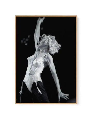 Framed black and white photograph of Madonna performing on stage, captured mid-movement with her right arm raised high and her left arm extended downwards. She is wearing a white conical bra corset with suspenders and dark pinstriped trousers, with a headset microphone. The background is dark, creating a dramatic contrast. The print is displayed in a natural oak frame with a white border.