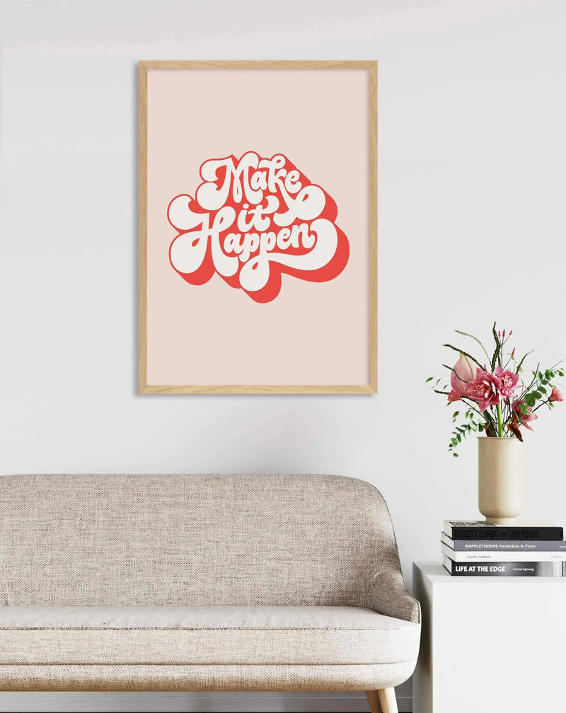 Framed art print displayed above a textured light beige sofa in a contemporary living room. The print features the words "Make it Happen" in a retro-inspired, bubbly red font with a white outline, set against a soft blush pink background, with a natural wood frame. To the right, a tall cream vase holds a bouquet of pink flowers and green foliage, resting on a stack of books on a white side table.