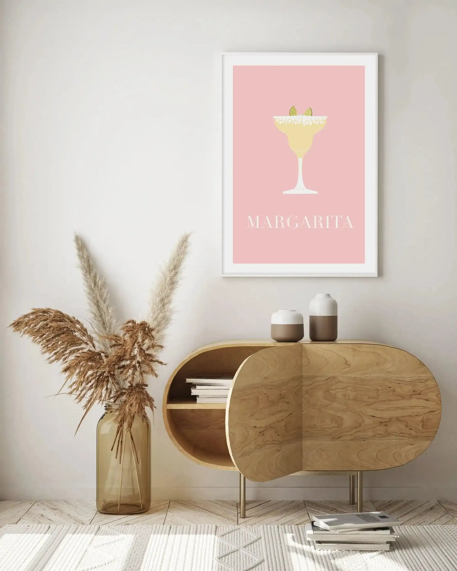 Lifestyle shot of a minimalist living space featuring a framed art print of a margarita cocktail on a light pink background. The print is displayed above a light wood, curved console table with two-toned ceramic vases and a stack of books. To the left, a tall amber glass vase holds dried pampas grass. The floor is light wood with a white textured rug.