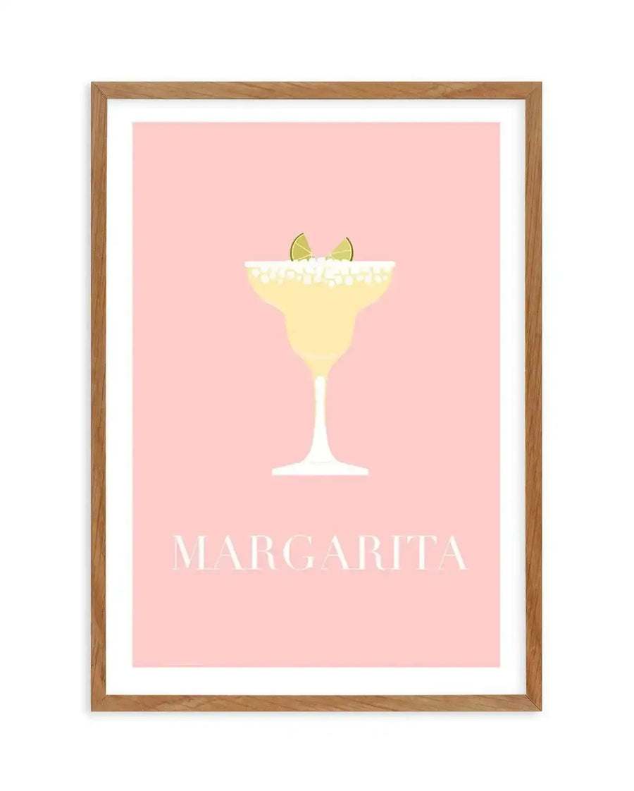 Art print featuring a minimalist illustration of a margarita cocktail in a classic glass with a salted rim and two lime wedges, set against a solid pastel pink background. The drink is a pale yellow colour, and the word "MARGARITA" is written in white capital letters at the bottom. The print is displayed in a natural oak frame with a white border.