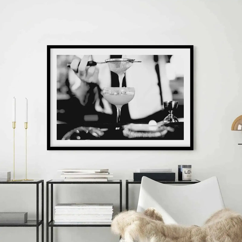Black and white photograph of a bartender's hands preparing a margarita cocktail, pouring the drink into a coupe glass with ice and a garnish, displayed in a black frame with white border on a light wall above minimalist furniture with books and gold candle holders.
