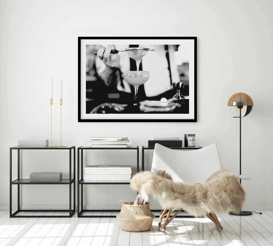 Lifestyle shot of a minimalist living room featuring a framed black and white art print of a bartender straining a margarita cocktail into a glass. The print has a black frame and white border and is displayed above a black metal console table with books and candles. A white armchair with a faux fur throw and a modern floor lamp are also visible.