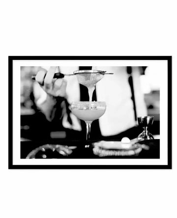 Black and white photograph in black frame with white border showing a bartender's hands straining a margarita cocktail through a fine mesh strainer into a coupe glass filled with ice. The bartender wears a white dress shirt and black vest. A small glass bottle and garnish visible on the dark bar counter in soft focus background. Professional, high-contrast photographic print capturing classic cocktail preparation technique.