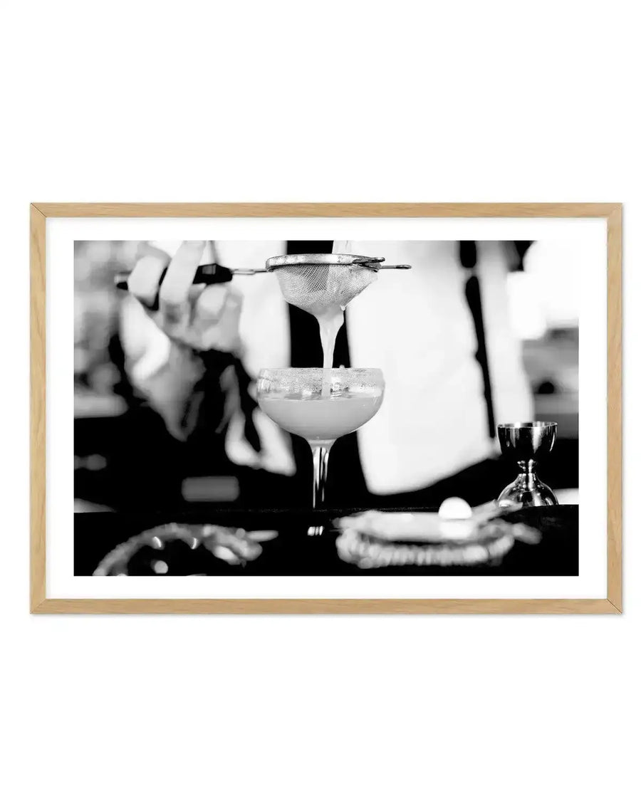 Black and white photograph of a bartender's hands preparing a margarita cocktail, straining liquid into a coupe glass with a salted rim. The image captures the classic cocktail-making technique with a bar strainer and jigger, with bottles and bar tools visible in the soft-focus background. Framed in natural wood with white border.