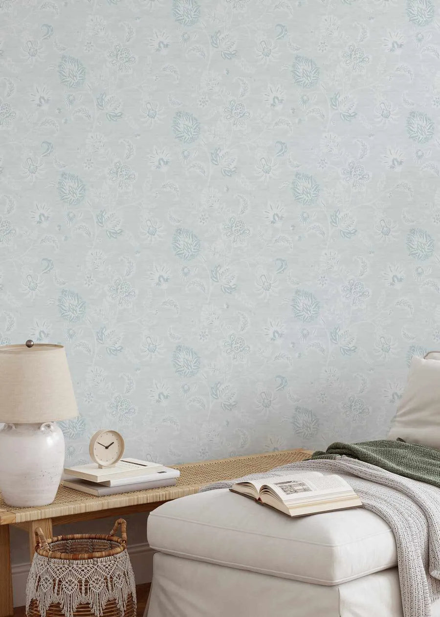 "Marsielle in Duck Egg Blue wallpaper featuring a repeating pattern of delicate, hand-drawn floral and botanical motifs in a soft duck egg blue and subtle white on a light grey-blue background, displayed on a wall in a bedroom. The room includes a light-coloured woven bench with a lamp, books, and a clock, and a white upholstered ottoman with a book and a green and grey throw."