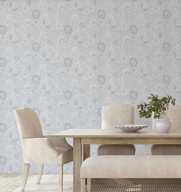 Lifestyle shot of a dining room featuring Marsielle in Salt wallpaper. The wallpaper has a repeating pattern of intricate floral and botanical motifs in a soft, muted grey and white on a light blue-grey background, creating a delicate, traditional damask-like design. A light wood dining table with two upholstered chairs and a bench in a neutral cream fabric are visible in the foreground. A white vase with green foliage and a decorative bowl sit on the table.