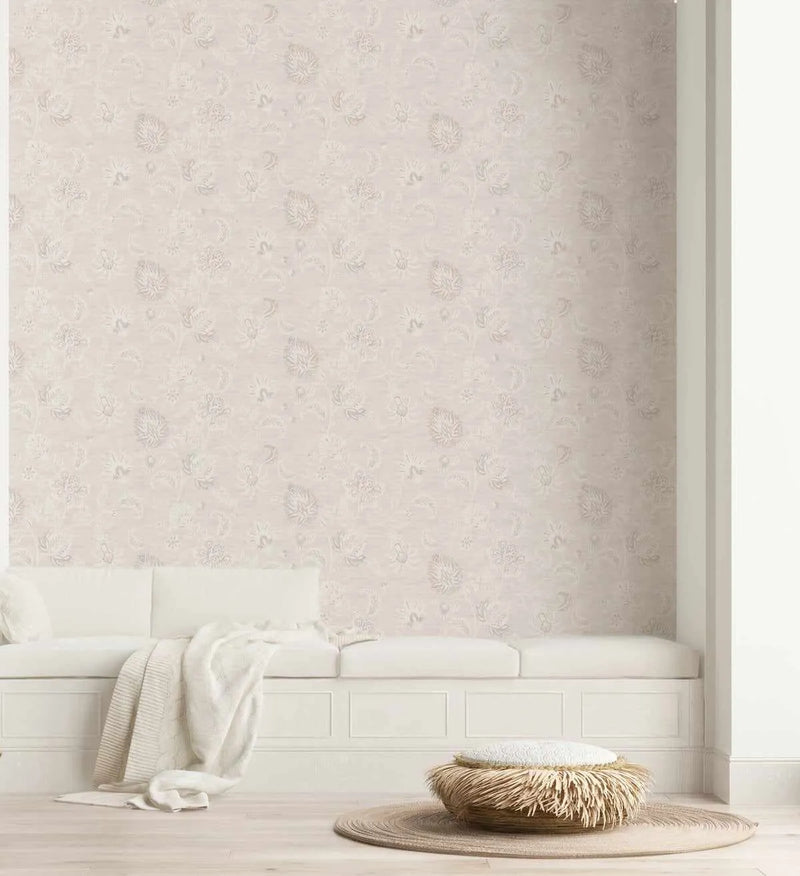 "Marsielle in Vanilla wallpaper featuring a repeating pattern of delicate floral and botanical motifs in subtle off-white and pale beige tones on a light cream background, applied to a wall above a built-in white bench seat with cushions and a cream throw. A round woven rug and textured ottoman are on the light wood floor."