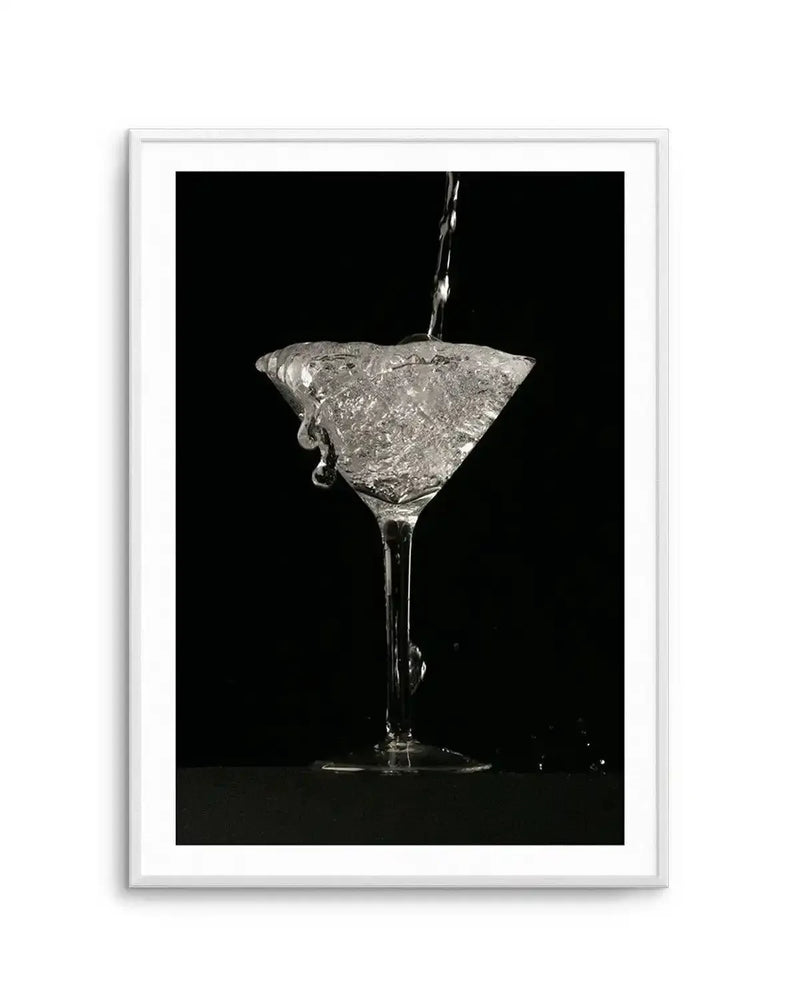 Black and white high-speed photography art print depicting a martini cocktail glass being filled with liquid against a solid black background. Water or clear liquid streams into the glass from above, creating dynamic splashes and droplets that freeze mid-motion around the rim and stem. The glass sits on a reflective surface with the stem clearly visible. The photograph captures the fluid movement and texture in stark monochromatic tones. The artwork is presented in a silver-toned frame with white border.
