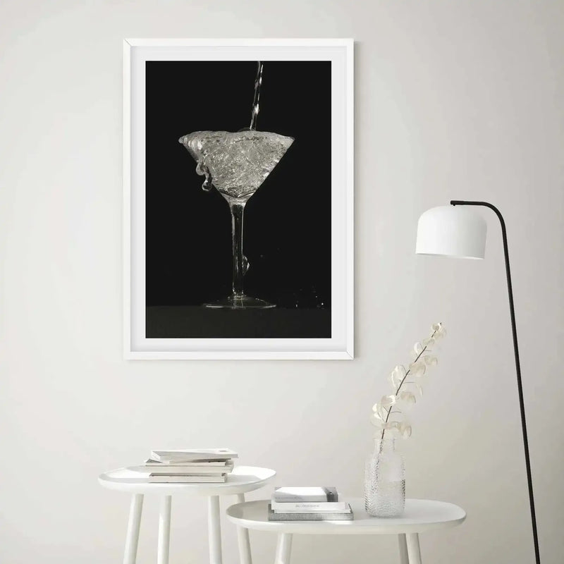 Black and white photographic art print in white frame with white border depicting a martini cocktail glass being filled with clear liquid against a dark background. The glass captures dynamic motion with splashing liquid and water droplets. Displayed on a white wall above a minimalist room setting with two small white round tables holding stacked books and a clear glass vase with dried white flowers, alongside a black arc floor lamp with white shade.