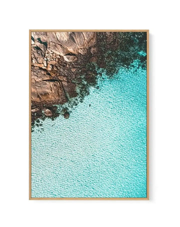 Aerial photograph framed in natural wood showing a rocky brown cliff face meeting turquoise water. The composition is split diagonally, with jagged tan and grey rock formations occupying the upper left portion, and calm, textured turquoise seawater filling the lower right. The photograph has a crisp, clear quality with visible water ripples and shadows in the rocks, conveying a coastal landscape viewed from above. The wooden frame has a warm, light natural finish.