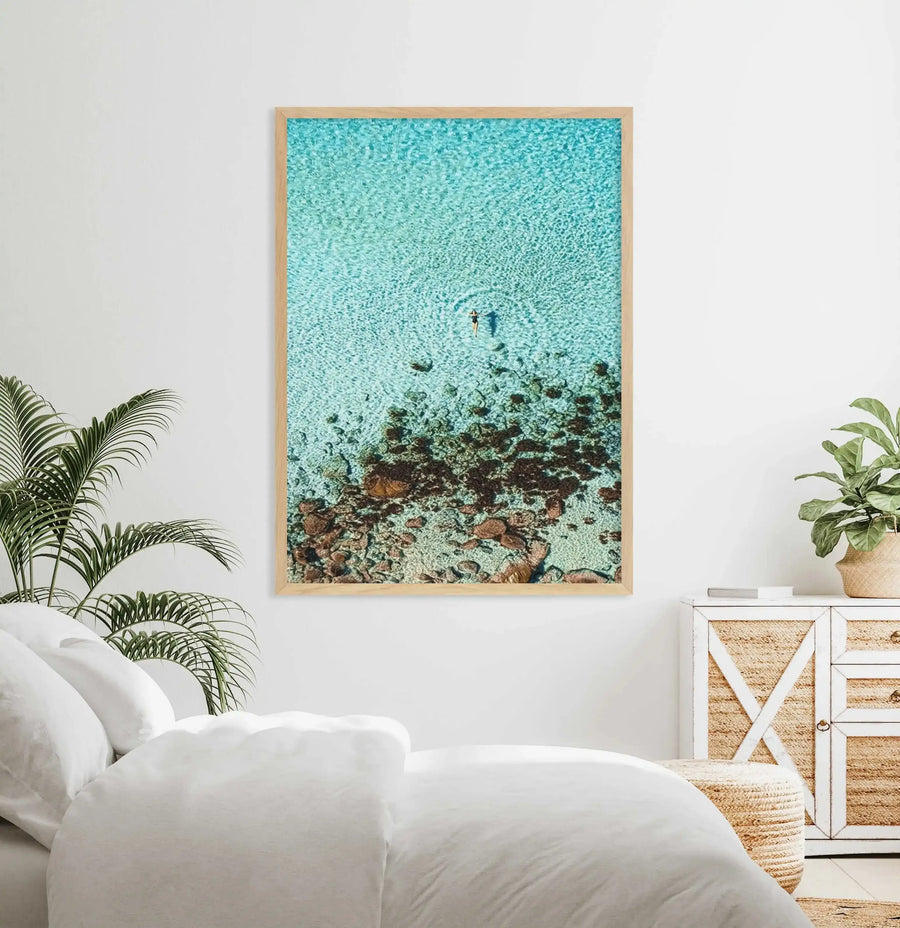 Lifestyle shot of a bedroom featuring a large framed aerial photograph of a person swimming in clear turquoise water with a rocky seabed, displayed above a white bed. The print has a natural wood frame and white border. The room includes a white duvet and pillows, a green potted plant on the left, and a white and woven cabinet with another potted plant on the right.